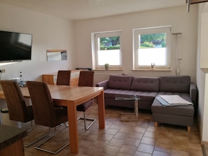Ein Wohnzimmer mit Sofa, Esstisch mit Stühlen, einem an der Wand montierten Fernseher und zwei Fenstern mit Jalousien.