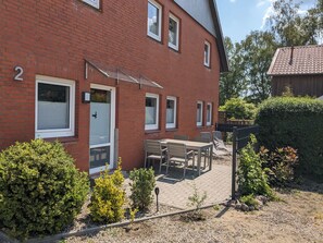 Ein rotes Backsteinhaus mit der Hausnummer 2, eine Terrasse mit Tisch und Stühlen und ein Hecke im Hintergrund.
