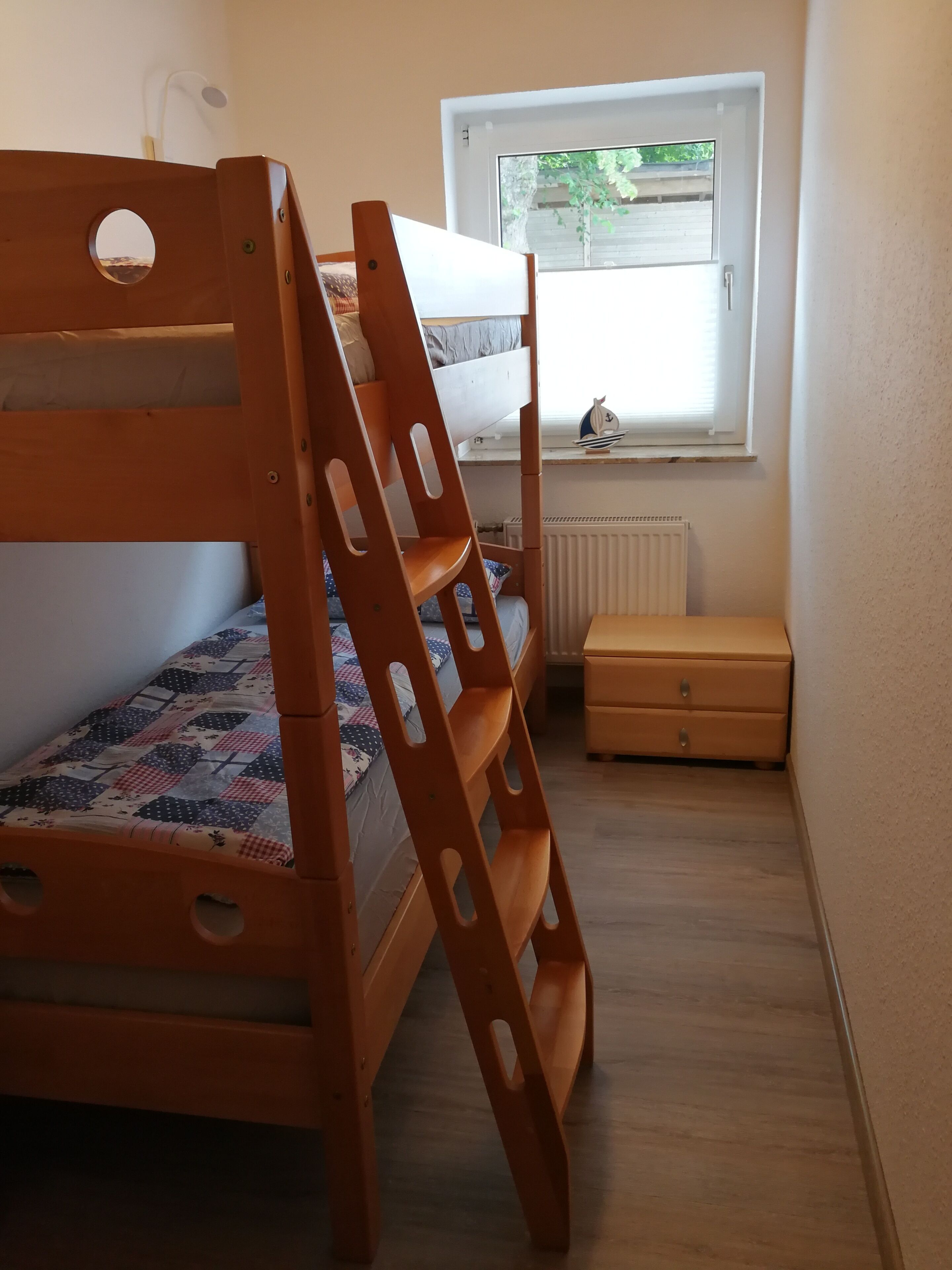 Una cama individual superpuesta con escalera, un pequeño cofre de madera y una ventana con vista a follaje.