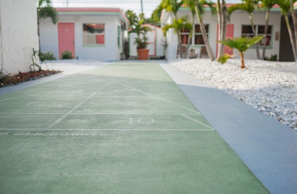 A green outdoor shuffleboard court with white lines, surrounded by a white gravel path and a row of potted plants.