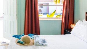A hotel room with a bed, a window with curtains, a hat, a towel, and a bottle of lotion.