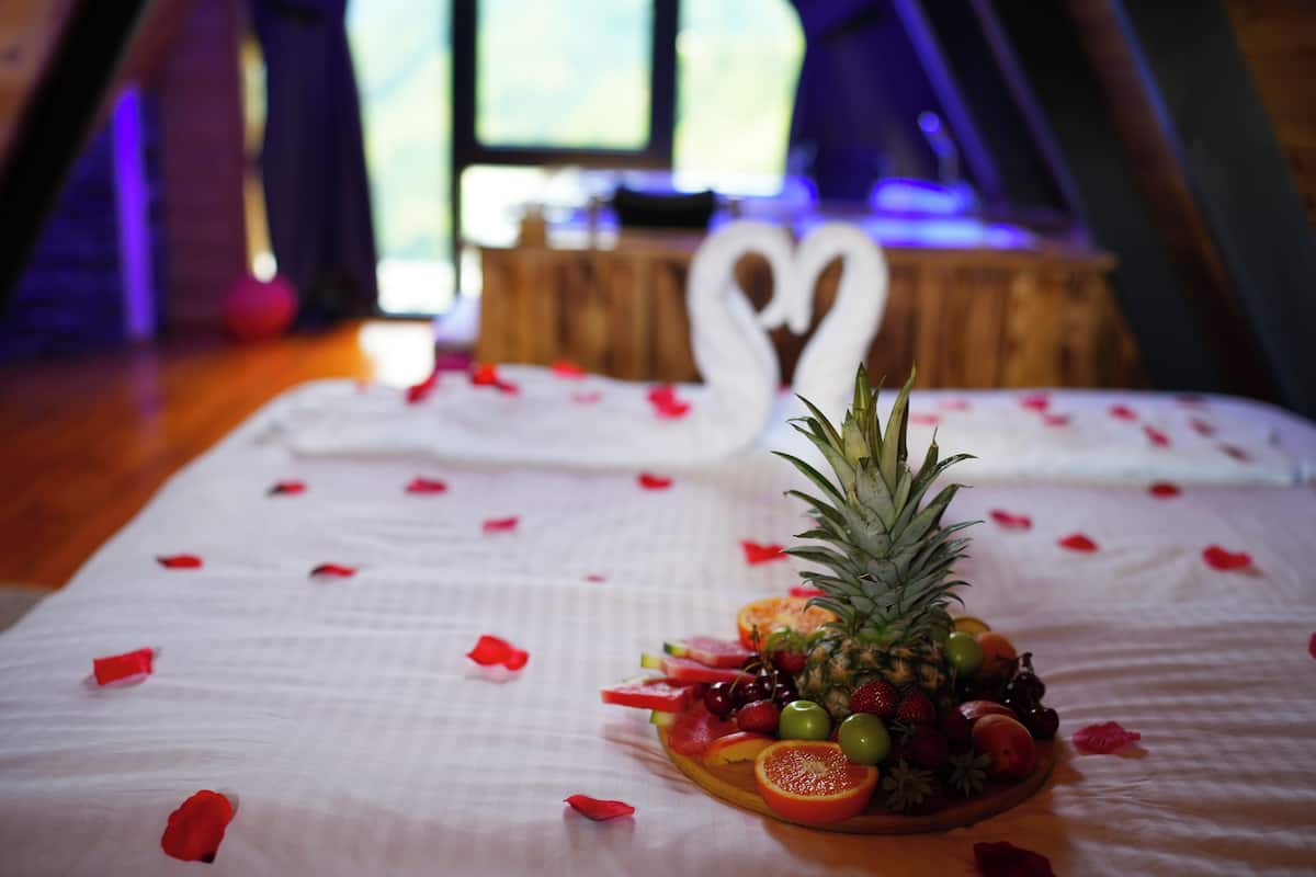 A bed with a fruit platter and rose petals, a white swan decoration, and a wooden table in the background.