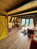 Una habitación estilo loft acogedora, con techo de madera, chimenea, sillón mecedora y un ventanal con vistas al exterior.
