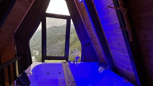A wooden cabin with a sloped ceiling and a large window. Inside, there is a wooden bathtub with a built-in seat and a small table. - Puleri Dağ Evi (Camlihemsin)
