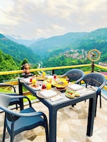 Una mesa preparada para el desayuno con vistas a la montaña, sillas de mimbre y un carretón decorativo.