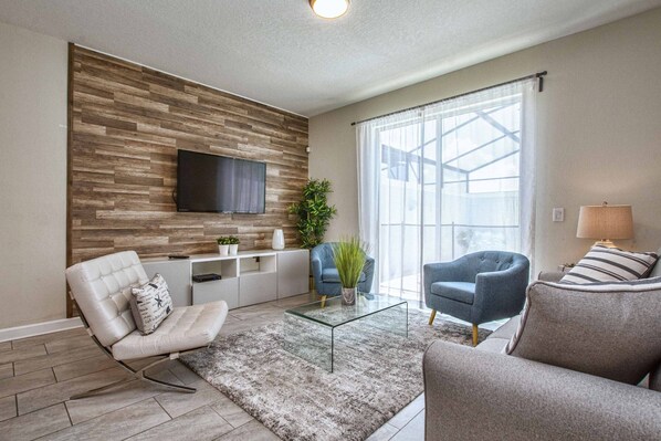 A modern living room with a flat-screen TV mounted on a wooden wall, a glass coffee table, and two sofas. - Disney Townhome | Waterfront Patio, Splash Pool, Waterpark, BBQ, Fast WiFi (Kissimmee)