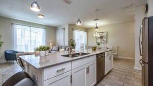 Une cuisine moderne dotée d’un îlot central, de meubles blancs, d’appareils électroménagers en acier inoxydable et de luminaires suspendus.