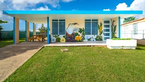 Een modern huis met een overdekte patio, een muurschildering van tropische vogels en bloemen, en een helderblauwe lucht.