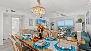Un coin repas avec une table en bois dressée pour quatre personnes, un lustre et une vue sur l’océan grâce à de grandes fenêtres.