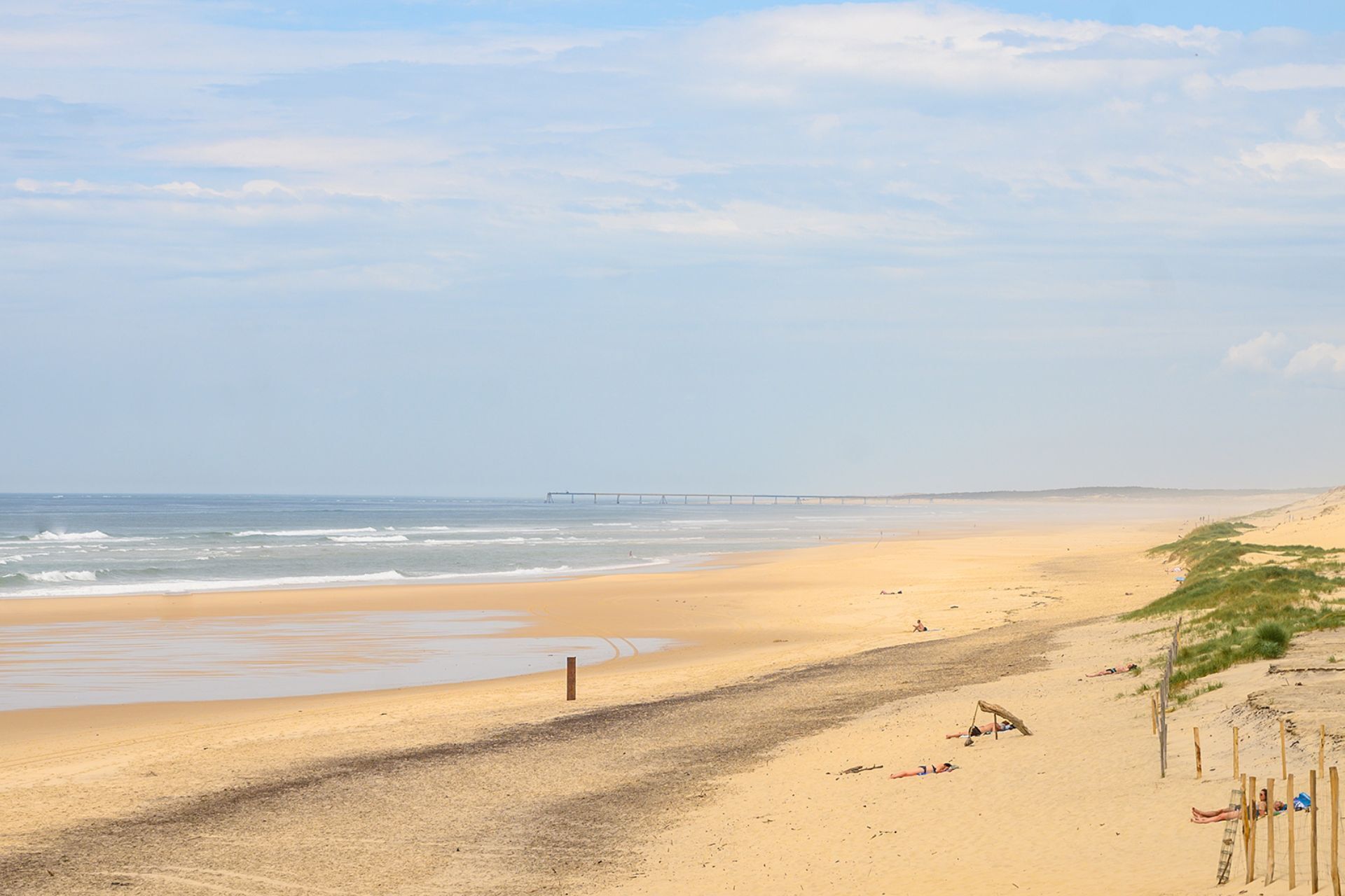 A beach with a long pier extending into the sea, sandy shores, and sparse vegetation.