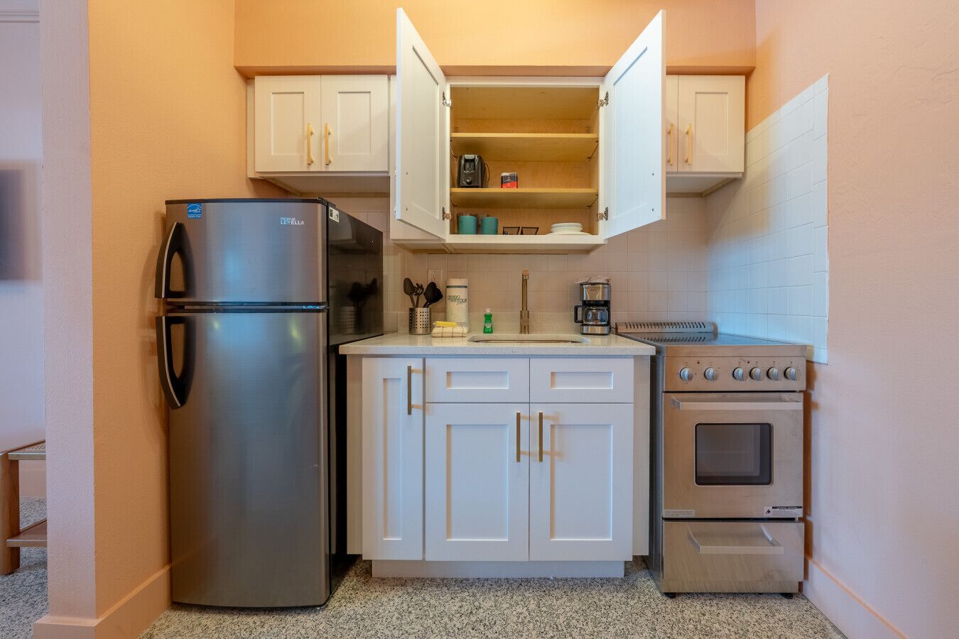 A compact kitchenette with a stainless steel refrigerator, a built-in oven, and a coffee maker.