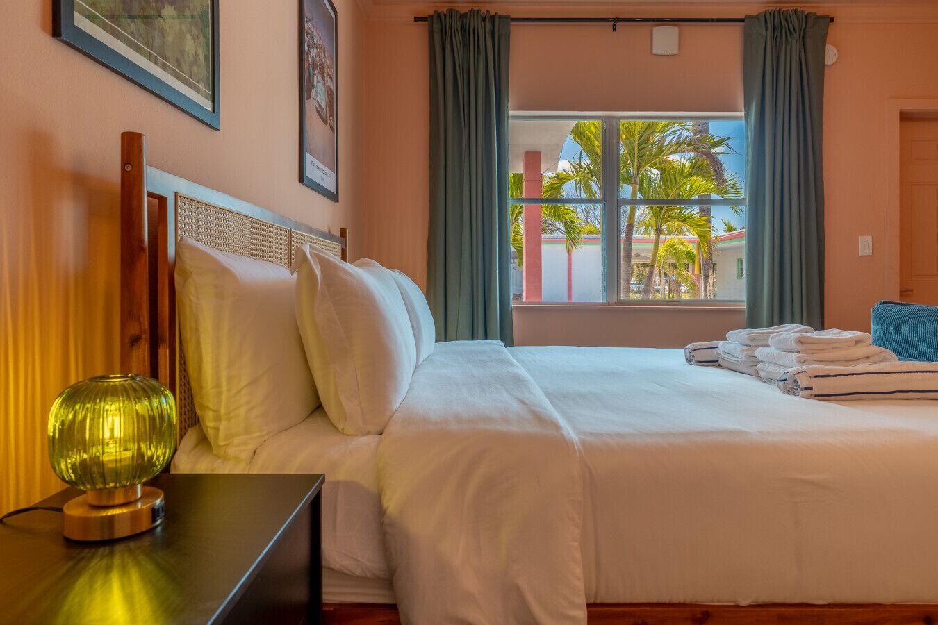 A hotel room with a bed, pillows, a bedside table with a lamp, a window with curtains, and a view of palm trees outside.