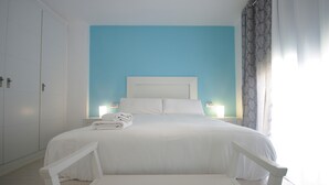 A neatly made bed with white linens and pillows, two bedside tables with lamps, and a view of a window with curtains. - 40 - Apartamento Sugar (Tarifa)