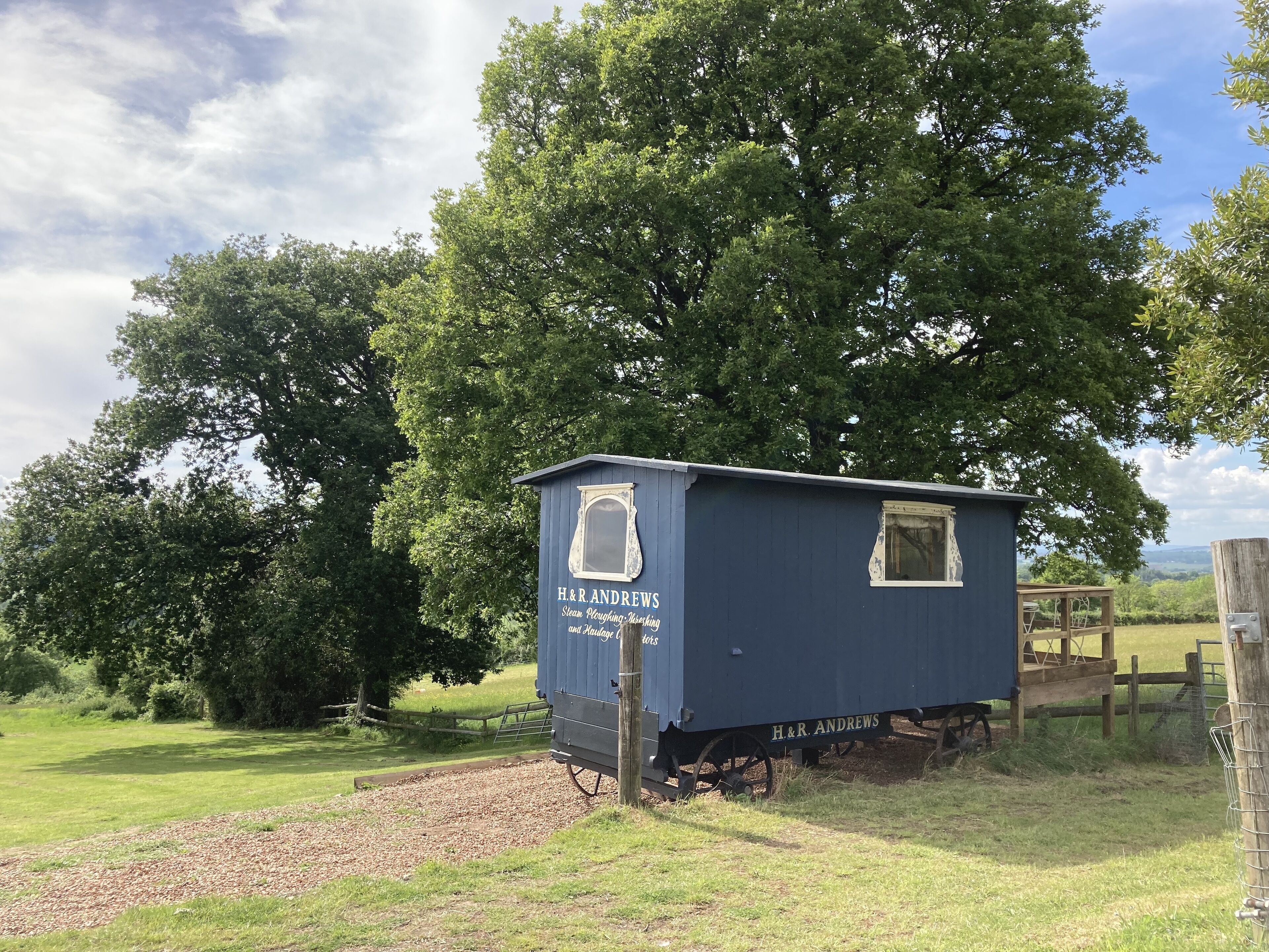 A small, single-story building with a dark blue exterior and a white-framed window. The building is labeled "H.R. ANDREWS Steam Puddling and Moulding Works." It is situated in a rural setting with a large tree in the background and a grassy area in the foreground.