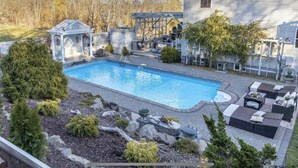 A rectangular swimming pool with a small gazebo, surrounded by a landscaped garden with rocks and shrubs, and a patio area with outdoor furniture.
