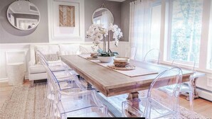A dining area with a wooden table, clear chairs, a white sofa, and a vase of white flowers.