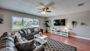A modern living room with a ceiling fan, a large TV, a sofa, a coffee table, and a patterned rug. - Pool - Hot Tub - BBQ - Private Backyard (Cape Coral)