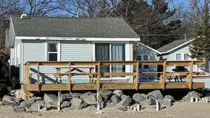 Une maison de plain-pied avec un toit gris et un revêtement extérieur blanc, une terrasse en bois à l’avant et une côte rocheuse à l’arrière.