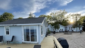 Une terrasse en bois avec des chaises longues et un espace barbecue couvert, donnant accès à une plage bordée d’une rangée de cabanes.