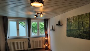 Et soveværelse med en seng, to vinduer, en radiator, en vægmonteret lampe og et billede med skovmotiv.