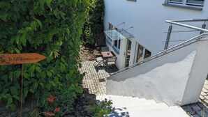 En trappe fører op til en bygning med et 'Willkommen'-skilt, et lille udendørs siddeområde og udsigt over det grønne omkringareal.