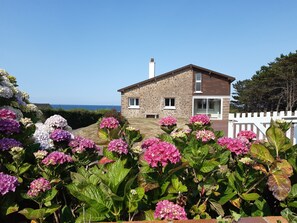 Une maison en pierre avec une clôture blanche, entourée de fleurs d’hortensia.