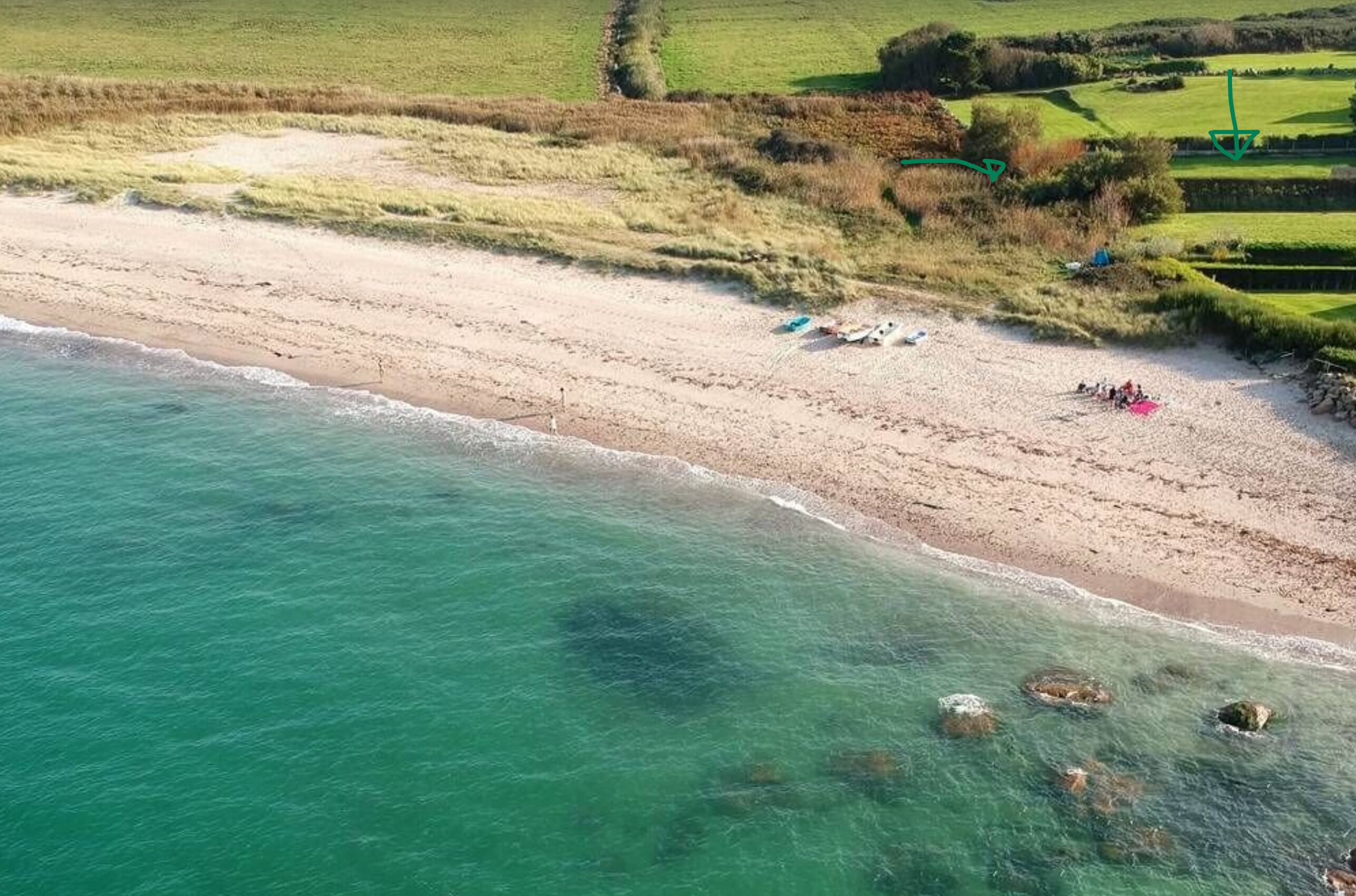 An aerial view of a beach with clear turquoise waters, a sandy shore, and some scattered rocks.
