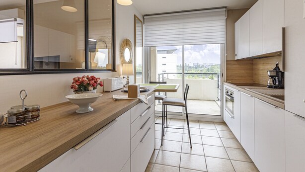 Une cuisine moderne avec des armoires blanches, un comptoir en bois, un coin repas comprenant une table et des chaises, et une grande fenêtre offrant une vue sur la ville.