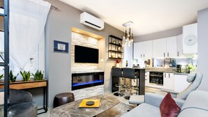 Una sala de estar moderna con chimenea de piedra, un comedor con mesa de mármol y una cocina con armarios blancos y un reloj.