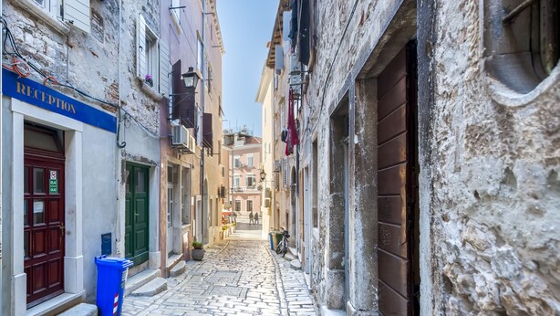 Una estrecha calle adoquinada flanqueada por edificios, un cartel de recepción y un bote de basura azul.