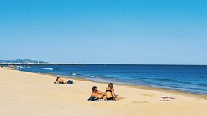 Une plage avec un ciel dégagé, un couple assis sur le sable et quelques personnes à l’arrière-plan.