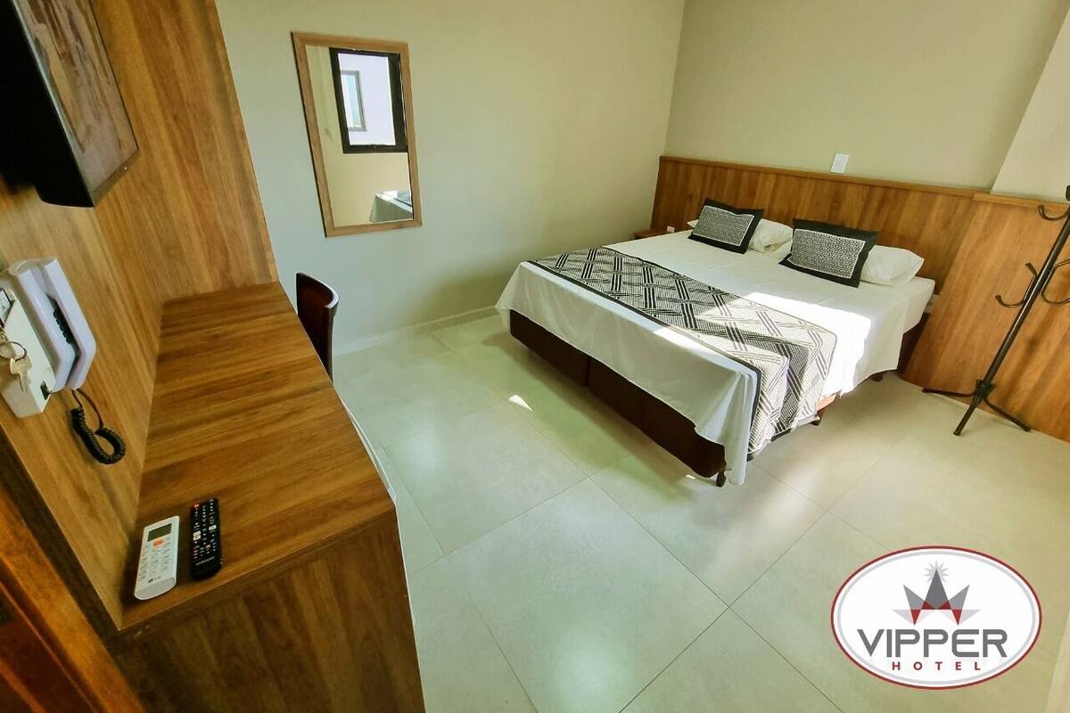 Une chambre d’hôtel avec un lit, un bureau en bois muni d’un téléphone et d’une télécommande, un miroir et une fenêtre.