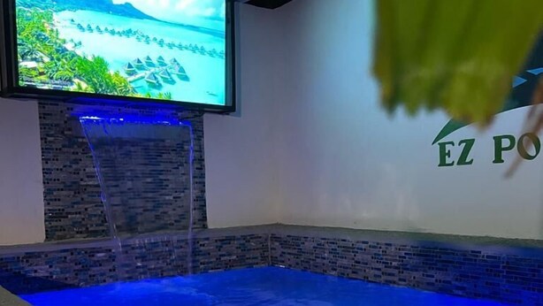 A pool area with a waterfall feature, a television mounted on the wall, and a logo on the wall. - Yellow Royal Family Suit 2brs, 3beds, 2bths, Pool (Portmore)