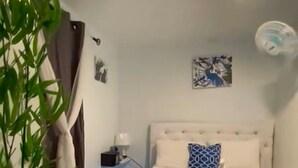 A bedroom with a bed, a nightstand, a lamp, a wall clock, a painting, a ceiling fan, and a geometric-patterned rug. - Yellow Royal Family Suit 2brs, 3beds, 2bths, Pool (Portmore)