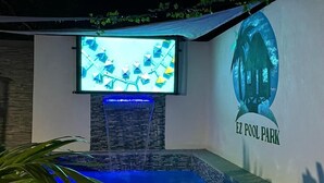 A swimming pool area with a waterfall feature, a mounted TV, and a mural of a house labeled "EZ POOL PARK". - Yellow Royal Family Suit 2brs, 3beds, 2bths, Pool (Portmore)