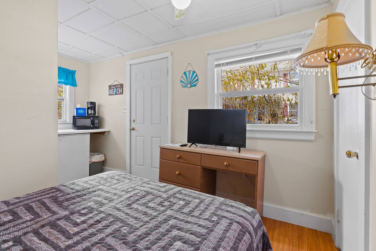 A bedroom with a bed, a desk with a television, a coffee maker, and a window with a view of trees.