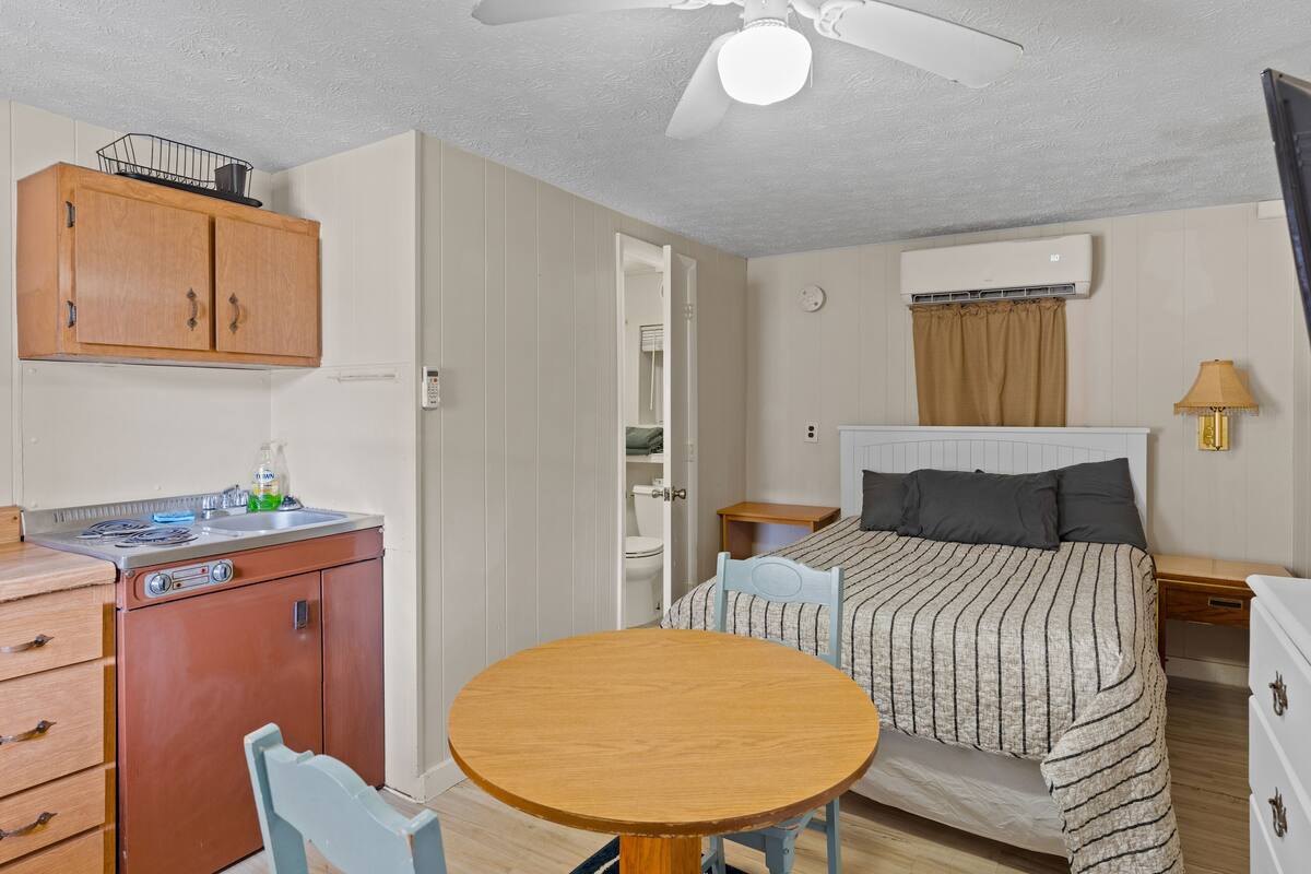 A compact room with a bed, a small table, a kitchenette with a fridge, and a ceiling fan.