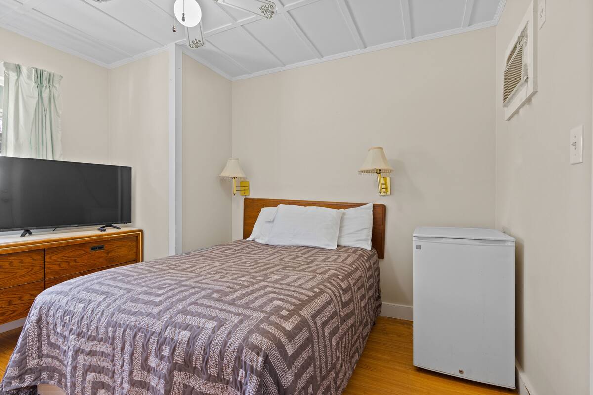 A bedroom with a bed, a television on a wooden dresser, two wall-mounted lamps, and a white refrigerator.