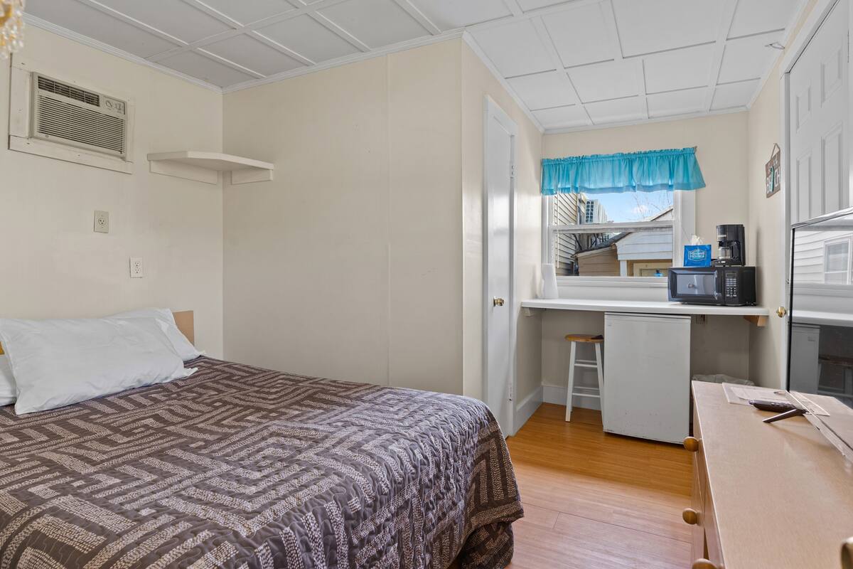 A bedroom with a bed, a desk, a kitchenette, and a window with a view of buildings.