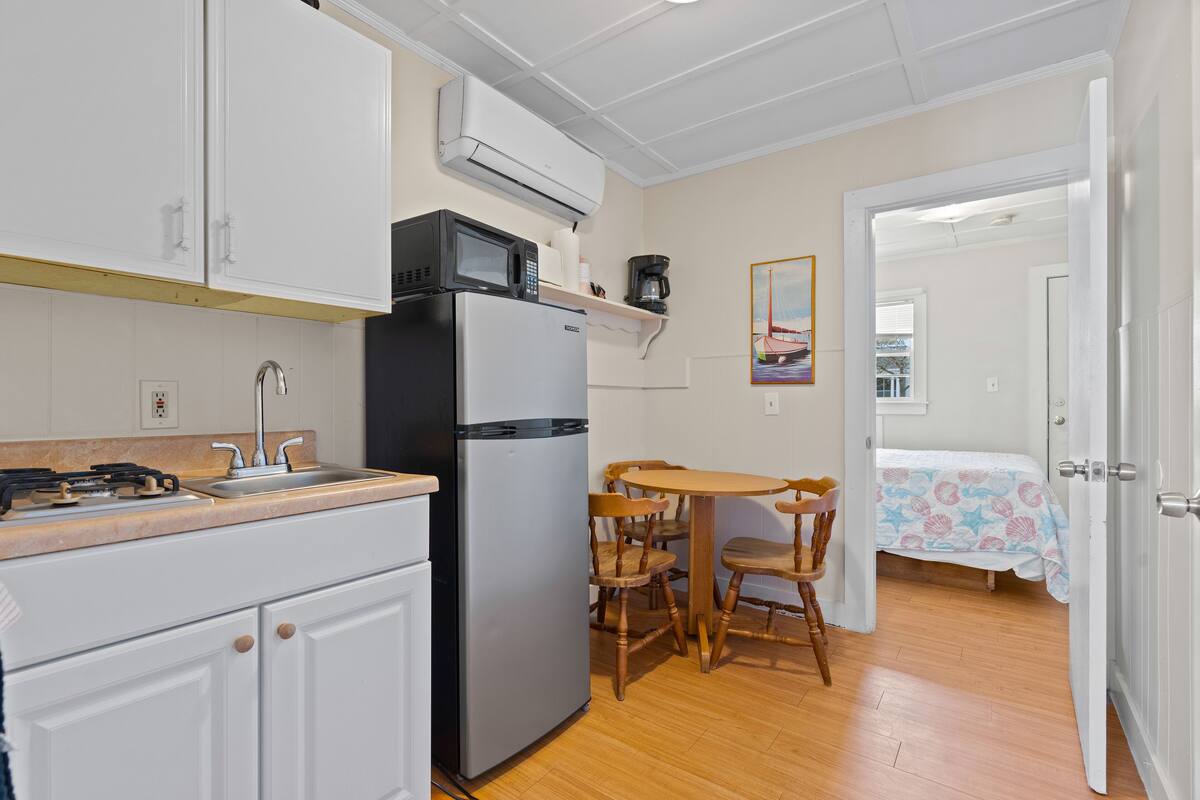A compact kitchen with a refrigerator, microwave, and stove. There is a dining area with a table and chairs. A bed is visible in the adjacent room.