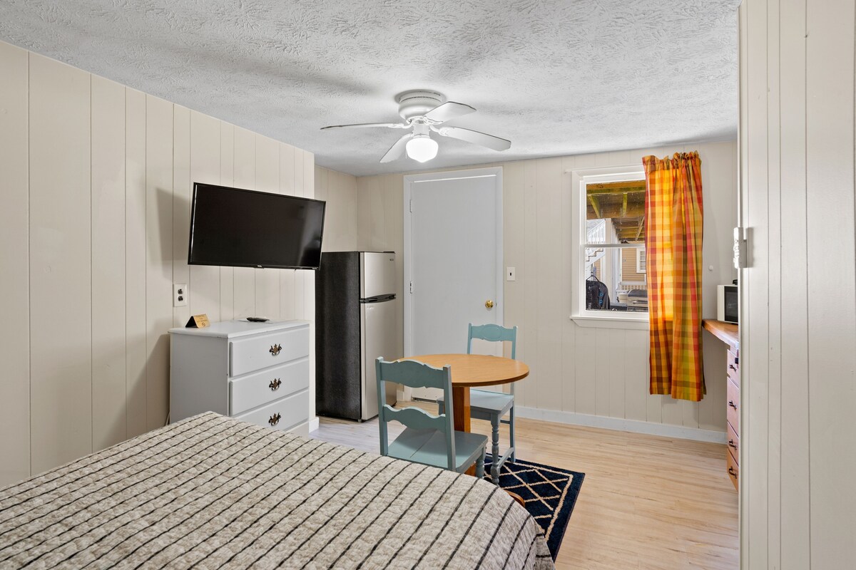 A bedroom with a bed, a television, a ceiling fan, a window with curtains, and a small table with chairs.