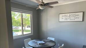 Un coin repas avec une table ronde, quatre chaises blanches, un ventilateur de plafond et une fenêtre donnant sur les voitures garées à l’extérieur.