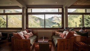 Una habitación acogedora con un gran ventanal que ofrece vistas a verdes colinas. Hay muebles de madera, incluyendo un sofá y una silla, y una pequeña mesa con un tablero de ajedrez.