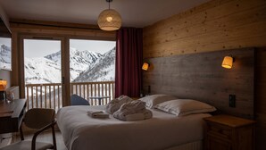 Un dormitorio con una cama grande, cabecera de madera, dos lámparas de noche, una mesita de noche de madera, una silla, un escritorio con lámpara y vistas a montañas nevadas a través de una puerta corredera.