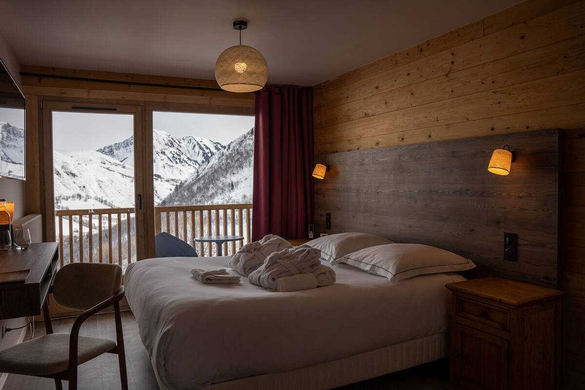 Un dormitorio con una cama grande, cabecera de madera, dos lámparas de noche, una mesita de noche de madera, una silla, un escritorio con lámpara y vistas a montañas nevadas a través de una puerta corredera.