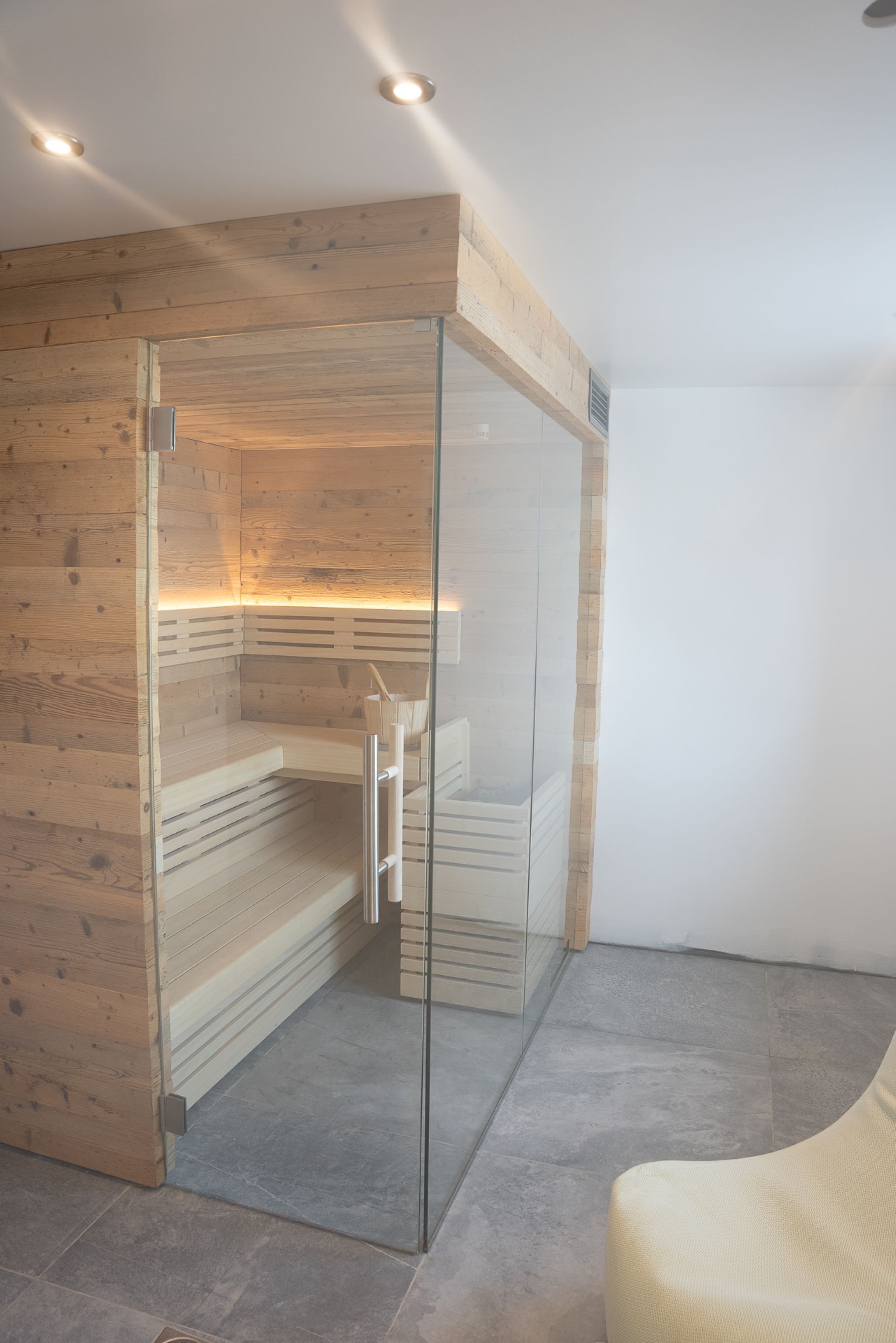 A modern spa room with wooden paneling, a glass-enclosed sauna, and a beige chair.