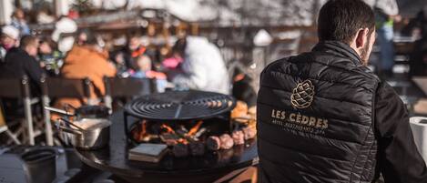 Eine Person mit Weste, auf der 'LES CÉDRES HOTEL RESTAURANTS SPA' steht, steht vor einem Grill, auf dem Essen zubereitet wird.