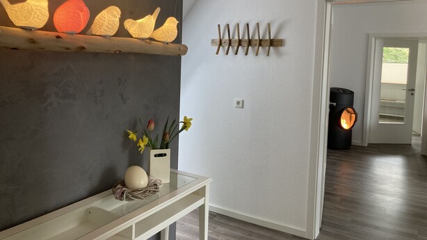 A modern interior with a dark wall, a wooden shelf with decorative lights, a glass table with a vase and flowers, and a fireplace. - Vacation cottage by the lake (Möhnesee)