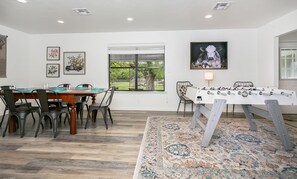 A dining area with a wooden table and chairs, a window with a view of greenery, and a foosball table.