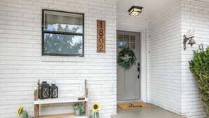 A white brick house with a wooden mailbox, a welcome wreath, and a bench with potted plants.